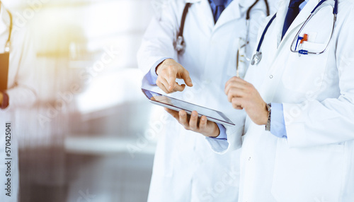 Group of unknown doctors use a computer tablet to check up some medical names records, while standing in a sunny hospital office. Physicians ready to examine and help patients