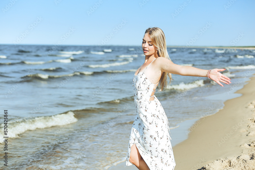 Happy blonde beautiful woman on the ocean beach standing in a white summer dress, open arms