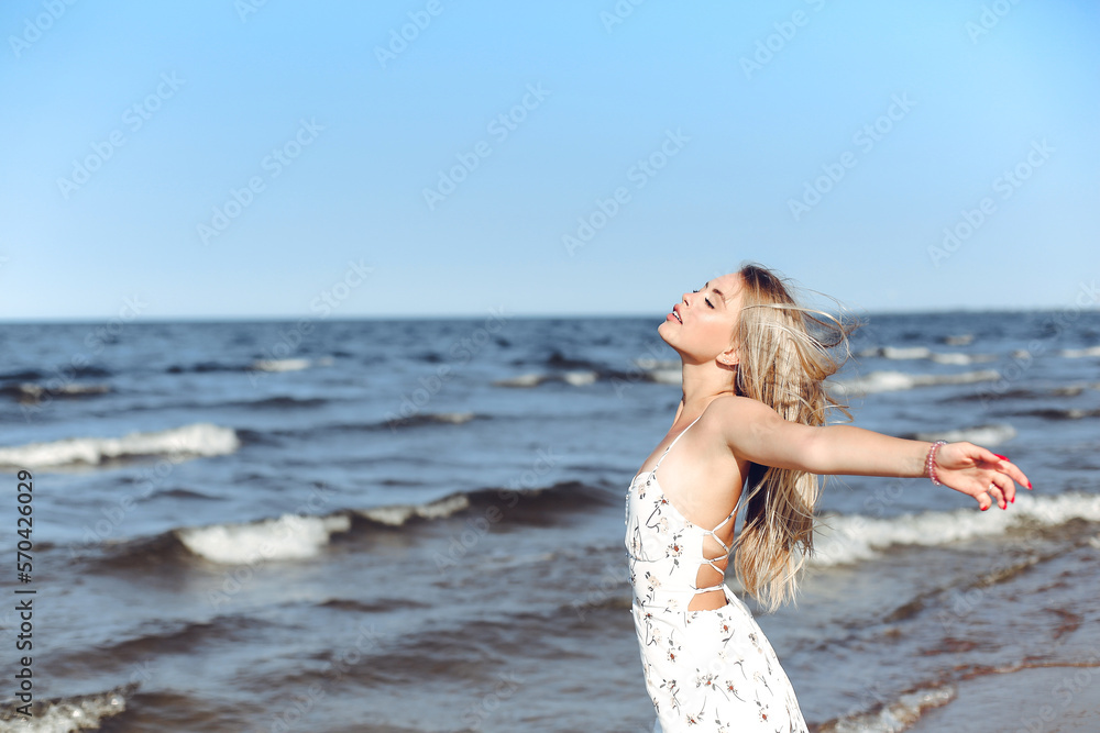 Happy blonde beautiful woman on the ocean beach standing in a white summer dress, open arms Happy blonde beautiful woman on the ocean beach standing in a white summer dress, open arms