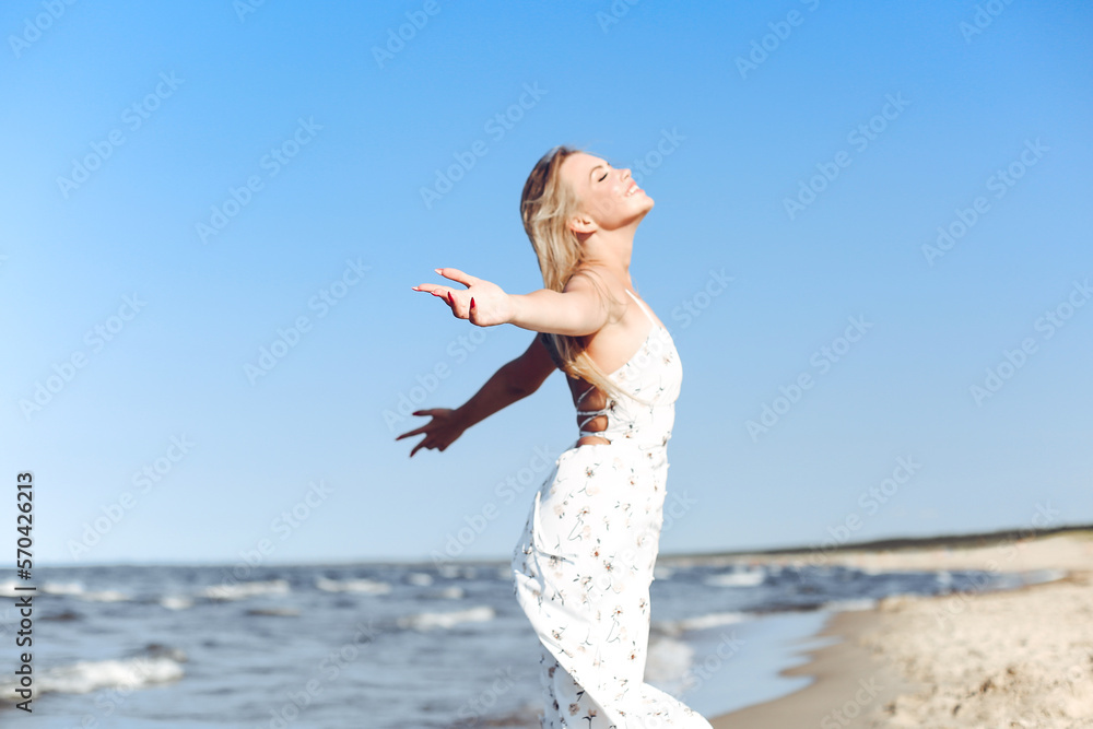 Happy blonde beautiful woman on the ocean beach standing in a white summer dress, raising hands Happy blonde beautiful woman on the ocean beach standing in a white summer dress, raising hands