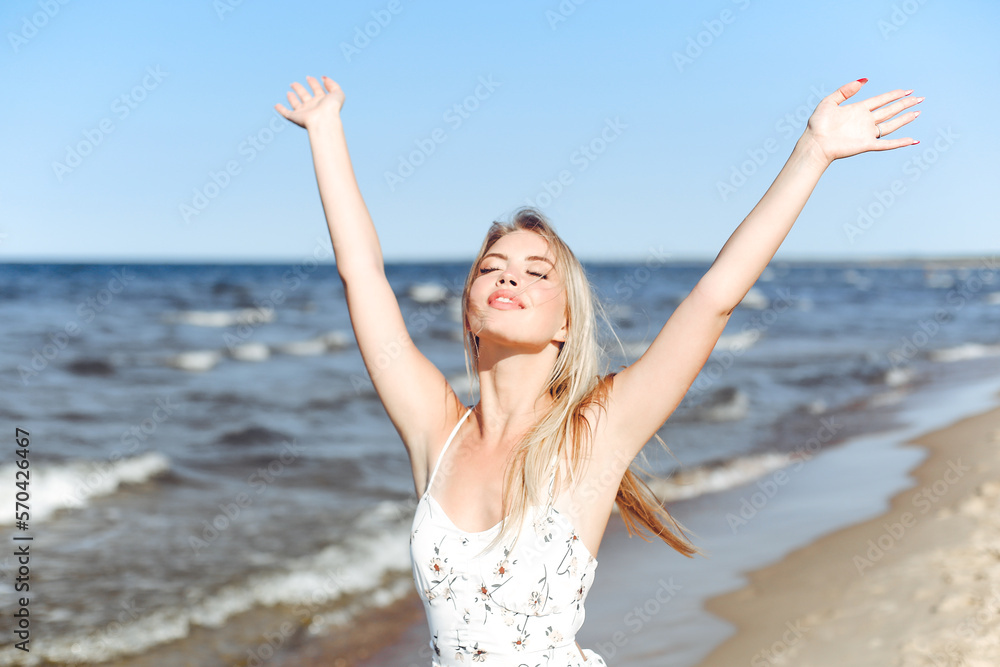 Happy blonde beautiful woman on the ocean beach standing in a white summer dress, raising hands Happy blonde beautiful woman on the ocean beach standing in a white summer dress, raising hands