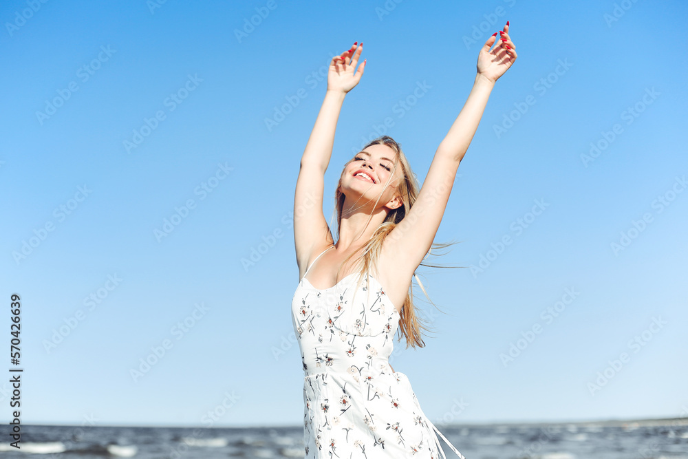 Happy blonde beautiful woman on the ocean beach standing in a white summer dress, raising hands