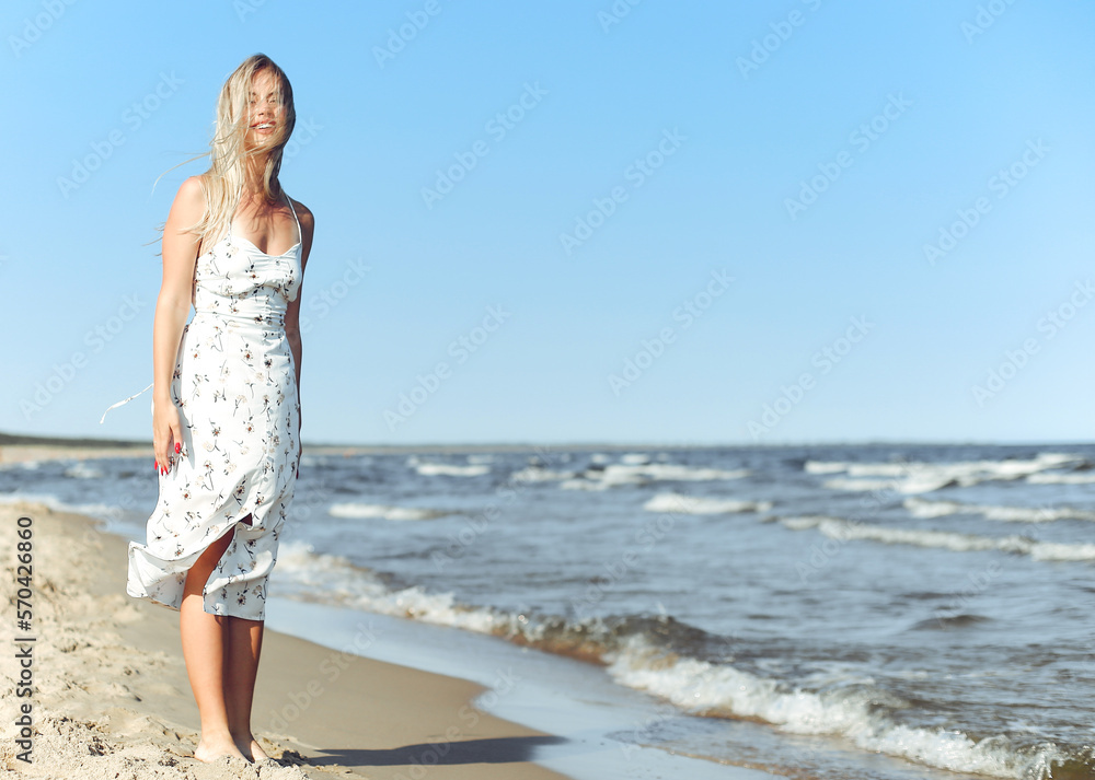 Happy blonde woman in free happiness bliss on ocean beach standing straight. Portrait of a female model in white summer dress enjoying nature during travel holidays vacation outdoors