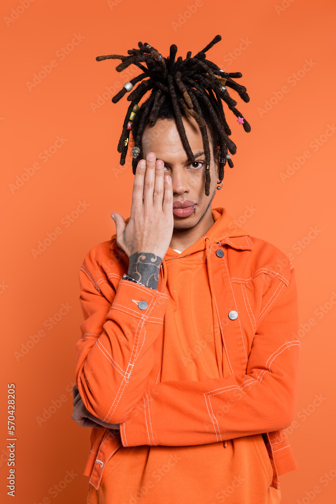 tattooed and multiracial man with dreadlocks covering half of face isolated on coral tattooed and multiracial man with dreadlocks covering half of face isolated on coral