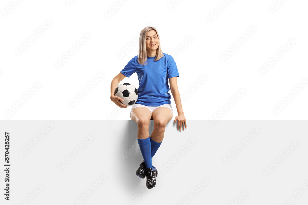 Full length portrait of a female football player sitting on a blank white panel and holding a ball Full length portrait of a female football player sitting on a blank white panel and holding a ball