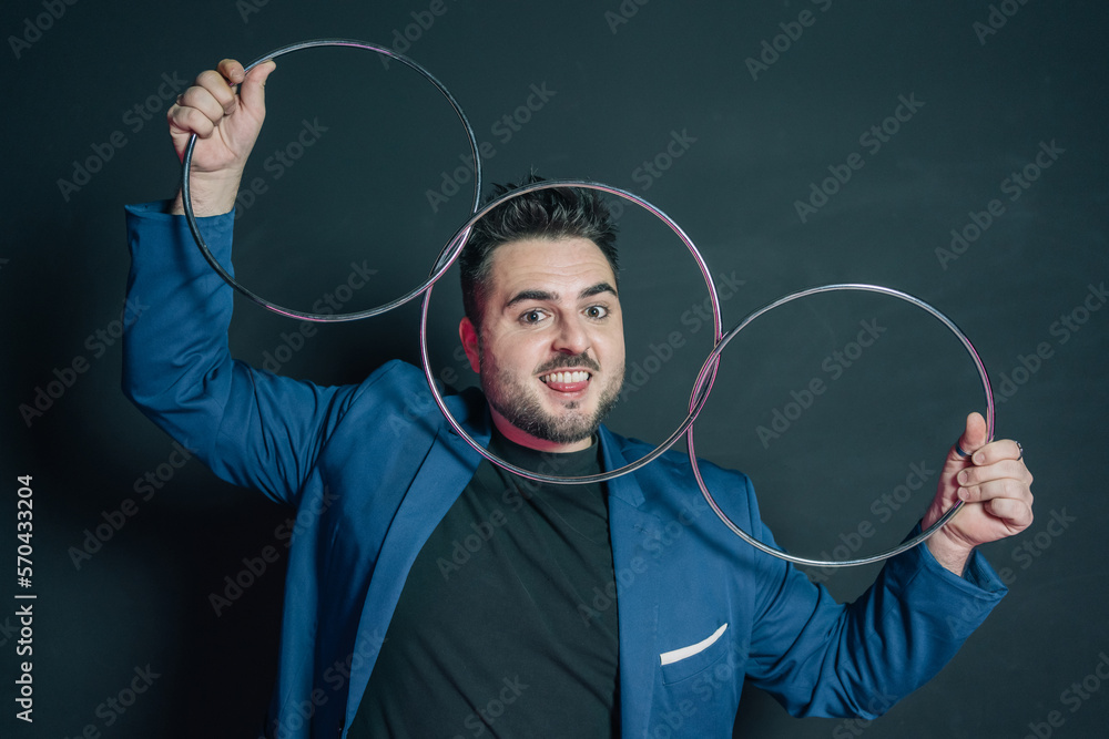 Young magician in the dark holding with both hands a set of three linked metal rings while looking at the camera and biting his tongue and placing his face just behind a ring.