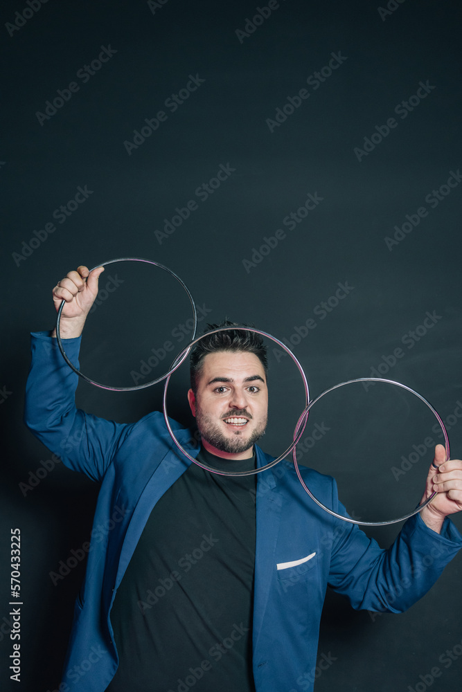 Vertical image of a young magician in the dark holding with both hands a set of three linked metal rings while looking at the camera and placing his face just behind one of the rings.