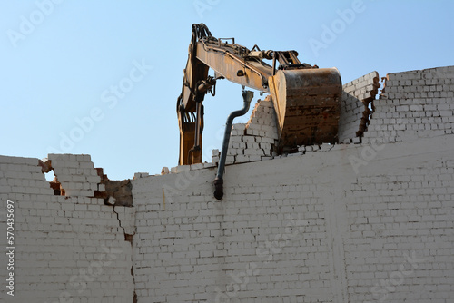 Фотография An excavator bucket is dismantling an old brick wall against the background of t