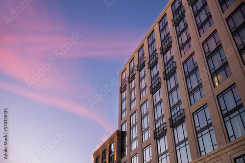 The facade of a building with one room illuminated by a table lamp, photographed at sunset with an afterglow of the clouds above in the sky.
