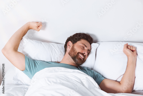 Happy man stretching on comfortable pillows in bed at home