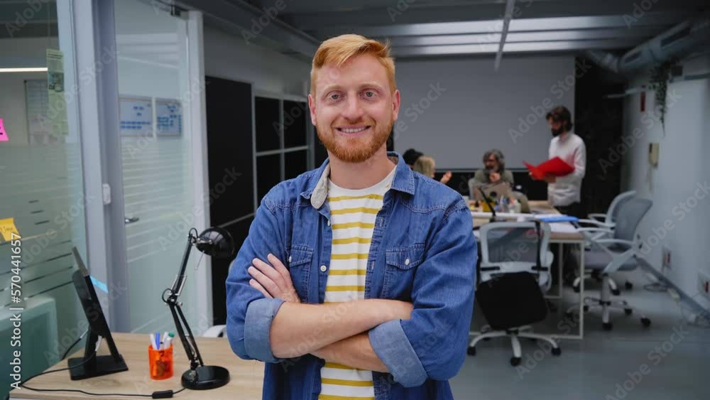 Young red-haired programmer or businessman in a modern office looking camera and smiles. Self-employed guy with his arms crossed poses for a video in the meeting room. Colleagues work in background. Young red-haired programmer or businessman in a modern office looking camera and smiles. Self-employed guy with his arms crossed poses for a video in the meeting room. Colleagues work in background.