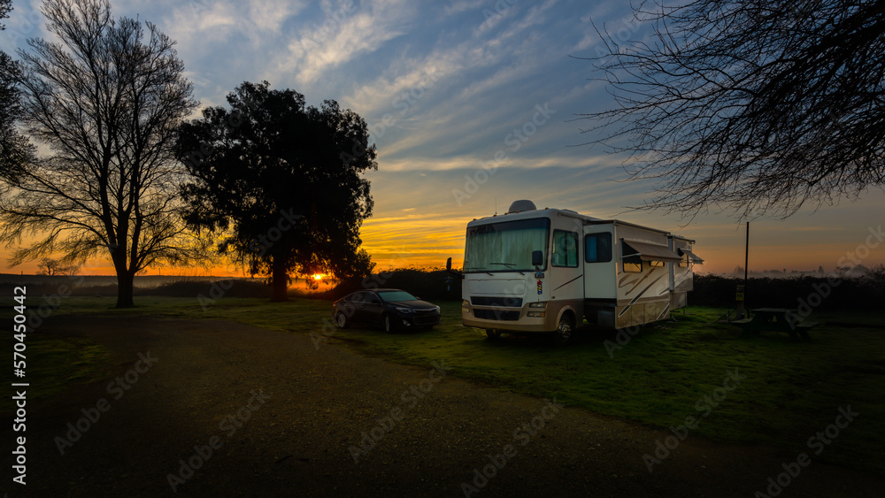 Rv motorhome and car parked under a rising sun beautiful blue sky and trees with cloud streaks
