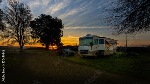 Rv motorhome and car parked under a rising sun beautiful blue sky and trees with cloud streaks 