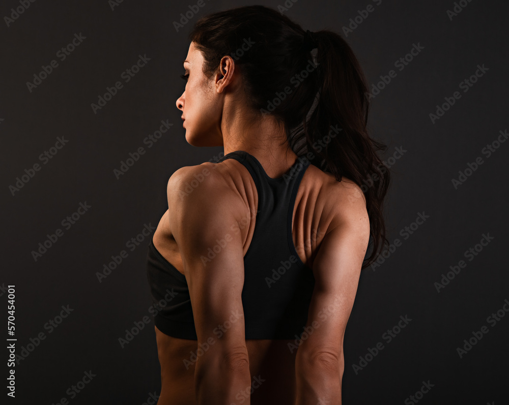 Serious female sporty muscular with ponytail doing stretching workout the shoulders, blades and arms in sport bra, standing on dark grey background with empty copy space. Back view. Lifestyle