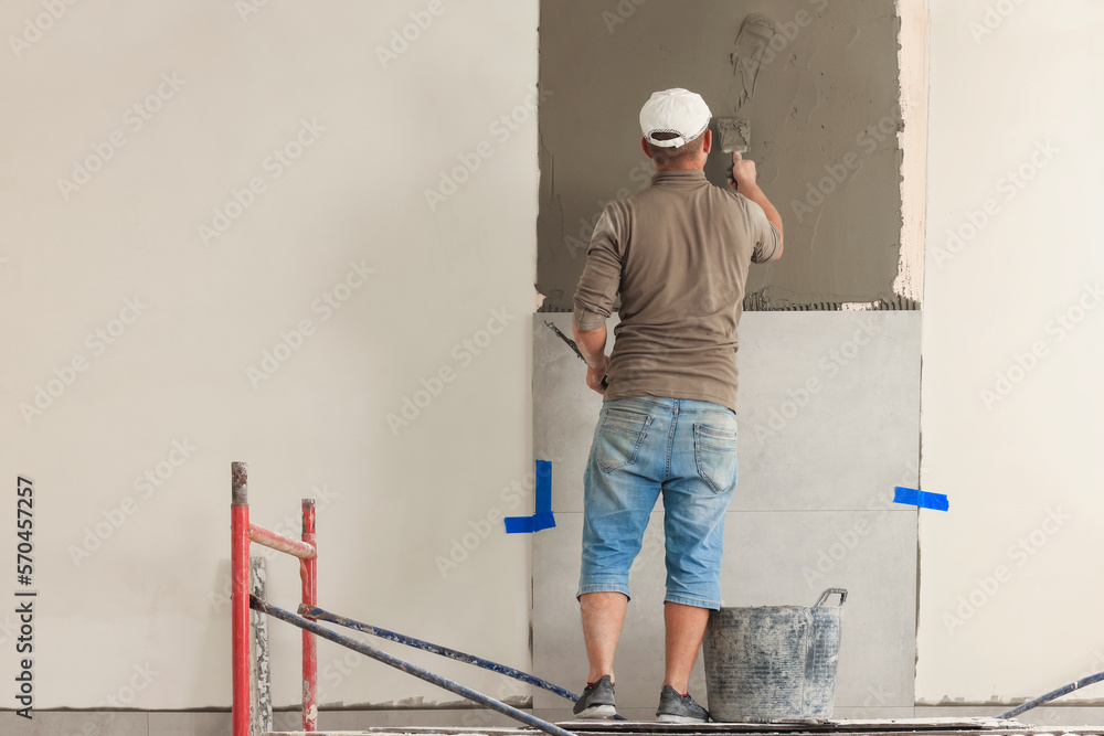 Worker spreading adhesive mix over wall for tile installation indoors, back view Worker spreading adhesive mix over wall for tile installation indoors, back view