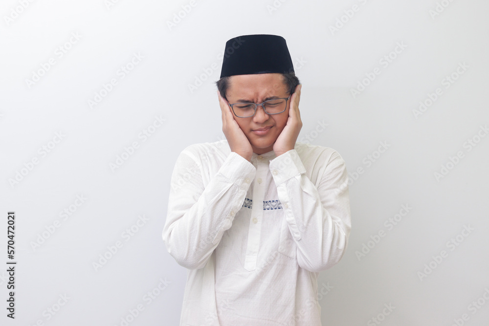 Portrait of young Asian muslim man covering ears with fingers with annoyed expression for the noise of loud music and sound. Isolated image on white background Portrait of young Asian muslim man covering ears with fingers with annoyed expression for the noise of loud music and sound. Isolated image on white background