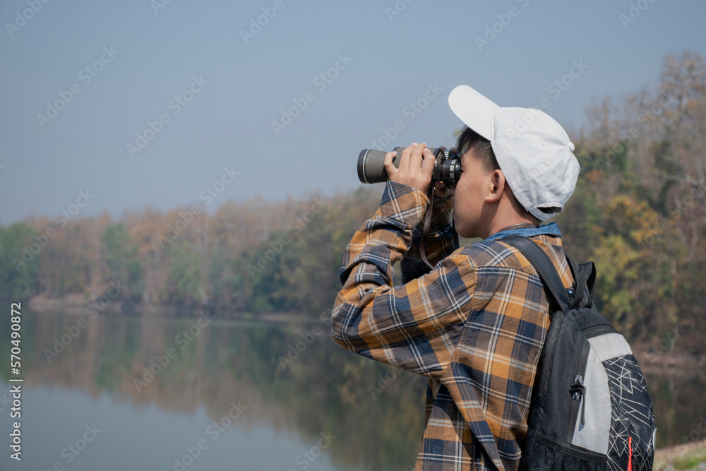 Asian boy in plaid shirt wears cap and has a backpack, holding a binoculars, standing on ridge reservoir in local national park to observe fish and birds on tree branches and on sky, soft focus.