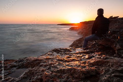 Man sitting on the rocks by the sea watching the sunset. Long exposure photo