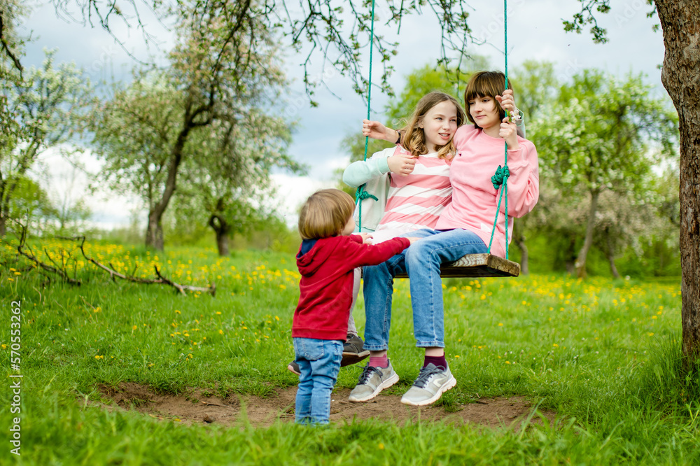 Two young sisters and their toddler brother having fun on a swing in blossoming apple orchard on warm spring day.