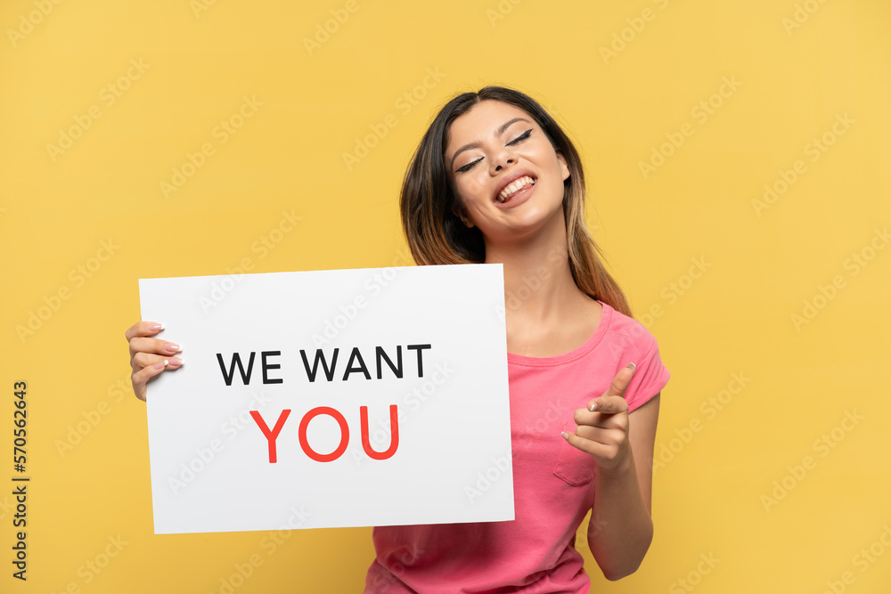 Young Russian girl isolated on yellow background holding We Want You board and pointing to the front Young Russian girl isolated on yellow background holding We Want You board and pointing to the front