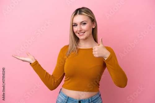Young caucasian woman isolated on pink background holding copyspace imaginary on the palm to insert an ad and with thumbs up