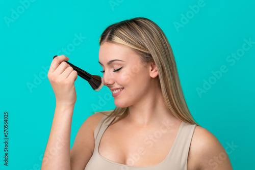 Young caucasian woman isolated on blue background holding makeup brush and whit happy expression