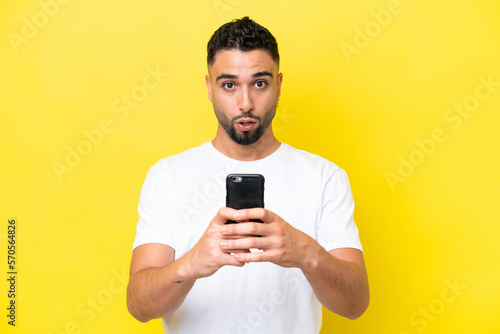 Young Arab handsome man isolated on yellow background looking at the camera while using the mobile with surprised expression © luismolinero
