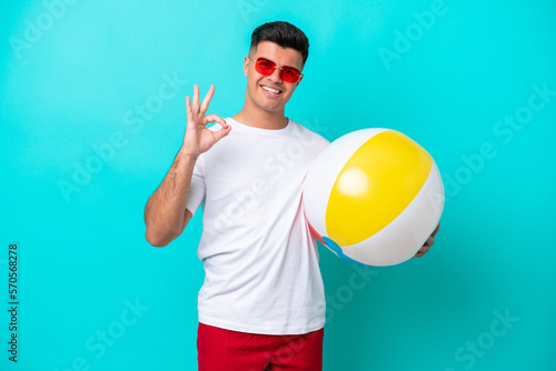 Young caucasian man holding a beach ball isolated on blue background showing ok sign with fingers