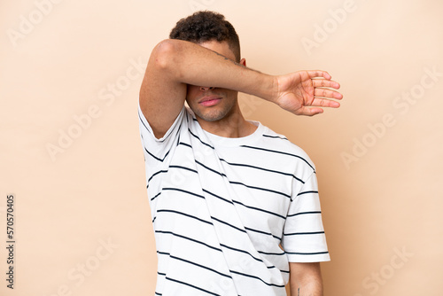 Young Brazilian man isolated on beige background covering eyes by hands