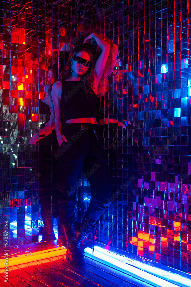 Fototapeta premium Portrait of a caucasian woman in sunglasses in neon light against a mirror wall. 