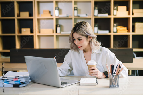 Confident beautiful Asian businesswoman typing laptop computer and digital tablet while holding coffee at office.