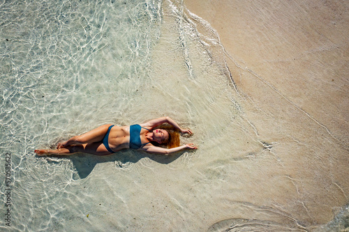 Fototapeta Naklejka Na Ścianę i Meble -  A young woman in a blue bikini lies on her back on the sand near the waves of the blue sea. View from above. The beach in the Maldives, clear clean water and yellow sand