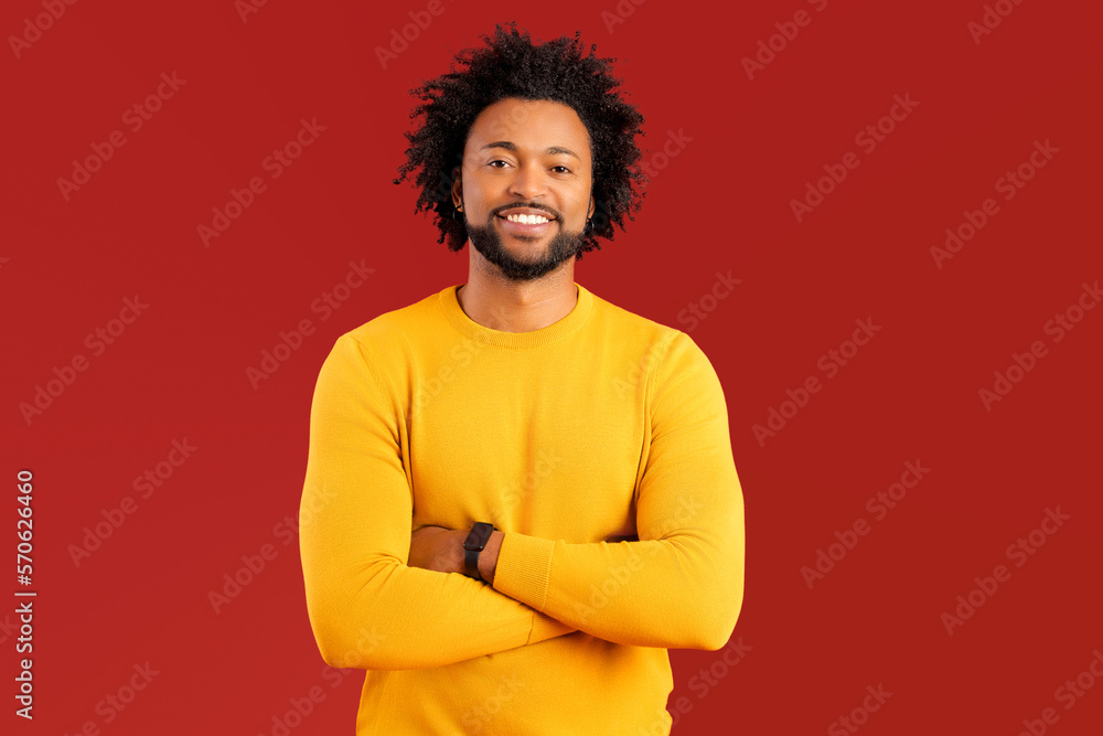 Handsome smiling African American curly young man in casual wear posing on red background, charismatic guy in yellow pullover with toothy smile stand with arms crossed and look at the camera Handsome smiling African American curly young man in casual wear posing on red background, charismatic guy in yellow pullover with toothy smile stand with arms crossed and look at the camera