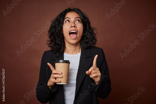 Young business woman drinking a take-away coffee pointing upside with opened mouth.