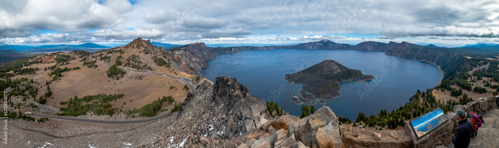 Panoramic view of Crater Lake with Wizard Island in the center and a sky with many clouds. The road that surrounds it and the viewpoint with some tourists can be seen in the foreground