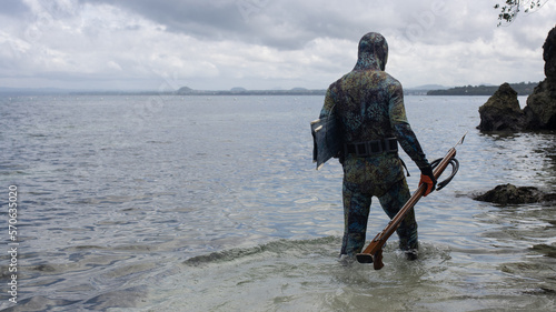 a man in a wetsuit, with camouflage coloring, is preparing for spearfishing, with an underwater rifle in his hands