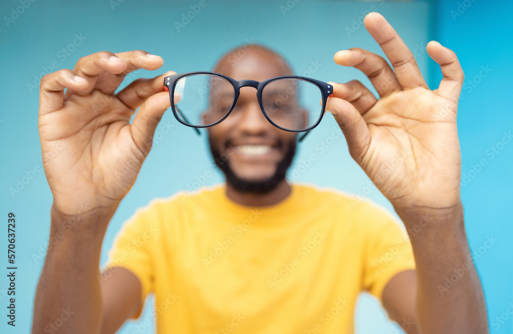 Fototapeta premium Hands, glasses and vision with a black man in studio on a blue background for prescription frame lenses. Spectacles, eyesight and eyewear with a male indoor to promote new round frames for seeing