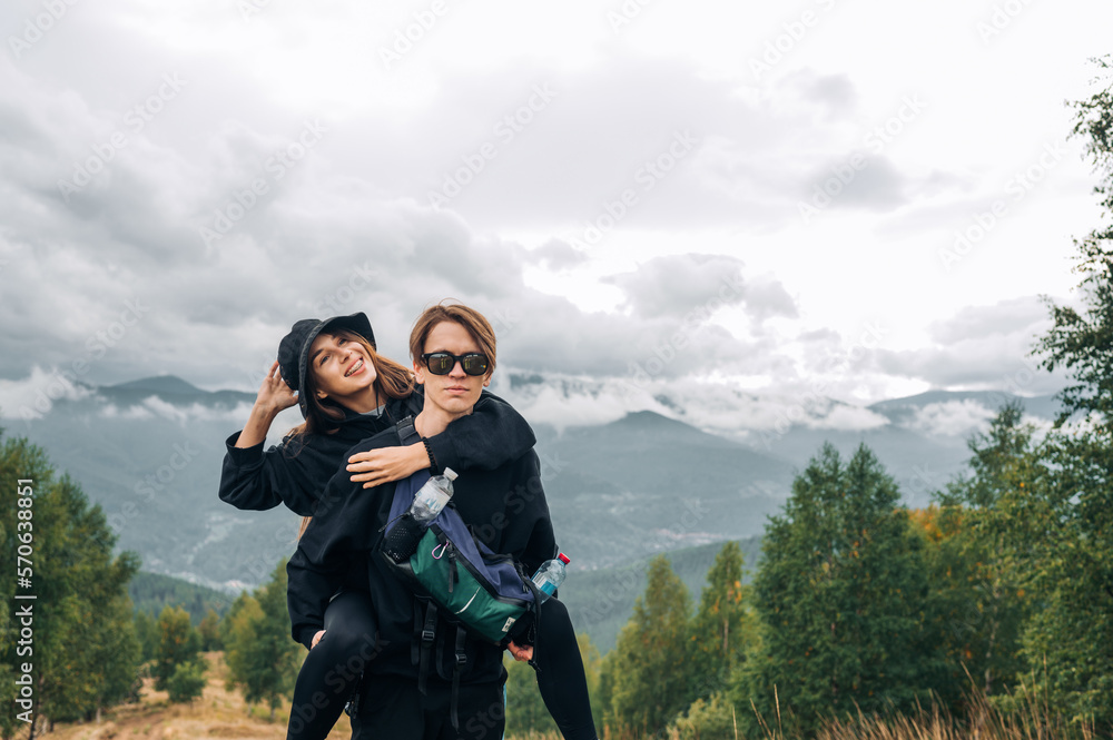 Positive hiking man and woman having fun after climbing a mountain, guy carries happy woman on back and looks at camera with serious face