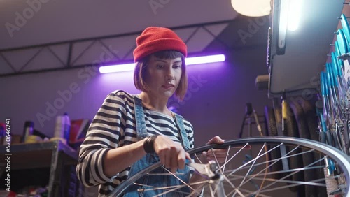 Side-view portrait of happy young caucasian female worker fixing bike wheel at workshop with tools. Beautiful hard-working woman mechanic repairing bicycles using instruments wrench to unscrew.