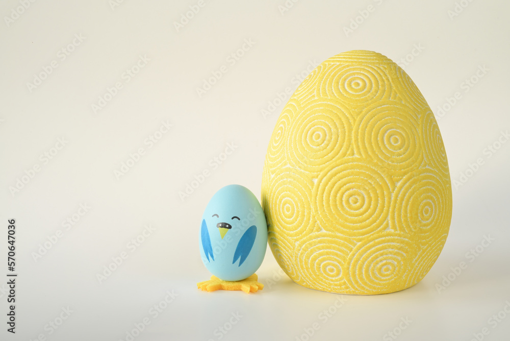 niebieski wielkanocny kurczak zrobiony z jajka stojący obok dużej żółtej pisanki na jasnym tle - obrazy, fototapety, plakaty 
