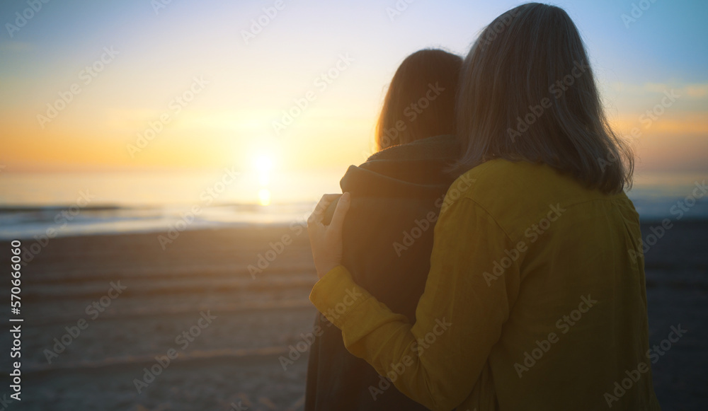 Mother cover his daughter with blanket and hugging on the beach at sunset.