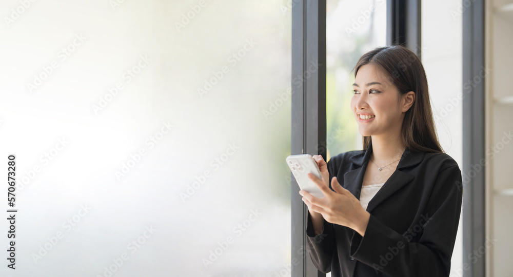 Portrait of elegant successful asian business woman speaking by smartphone and smiling happily while standing against window in office.
