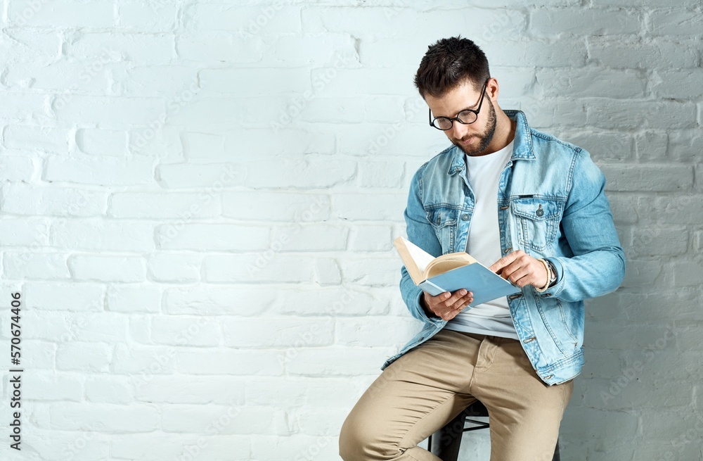 Young goodlooking caucasian man in glasses sitting at white brick wall, reading book, plenty of copyspace.