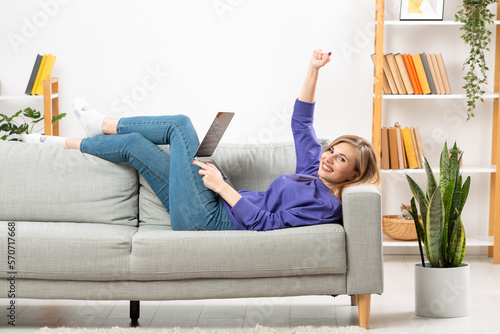 Portrait of an enthusiastic girl in full length, lying on the sofa, raising her fist, with a phone and a laptop