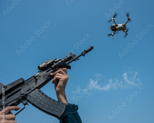 A man aims to shoot a rifle at a flying drone against a blue sky. 