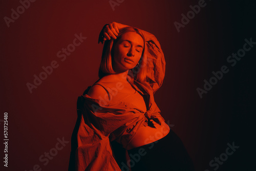 Relaxed blond lady in stylish outfit closing eyes and touching head while standing against red background in neon light