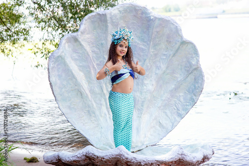 A cute girl in a mermaid costume stands outdoor in a large sea shell, holds her thumbs up