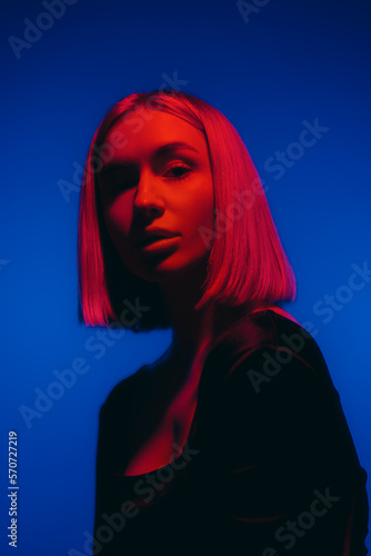 Young female with fair hair looking at camera against blue backdrop in studio with neon illumination