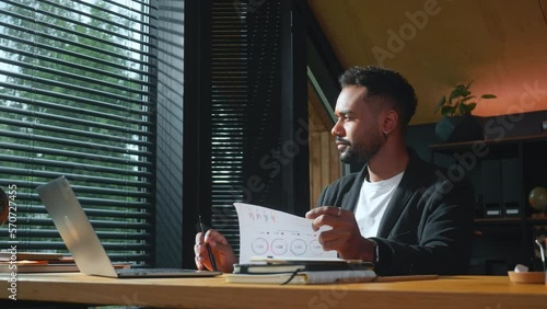 Happy smiling young finance expert businessman works at modern office table with documents, laptop looking at window. photo