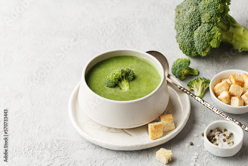 Green vegetable cream soup with broccoli and croutons in ceramic bowl over gray concrete background with ingredients. Side view